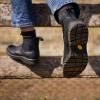 På billedet ses et par fødder klædt i robuste, sorte støvler med tykke, slidstærke såler. Støvlerne bæres af en person, der sidder med krydsede ben og hviler fødderne på en horisontal træbjælke. Støvlens design er karakteriseret ved en klassisk chelsea-boot silhuet med elastiske sider og en loopet trækhjælpeme på hælen for at hjælpe med at lette støvlerne på. De sortfarvede støvler kontrasterer med lysere blå denim jeans, som kun er synlige op til nedre del af kalven, hvor benet bøjes, og det indre frotté-forede materiale af sokken bliver synligt, mens den overlapper støvlens kant. Baggrunden er en sløret, naturlig scene, som indikerer, at billedet er taget udendørs, med hvad der ser ud som et hegn lavet af sammenflettet metal, der indrammer fokus på støvlerne. En orange firkantet etiket på den ene støvles sål giver et pop af farve og det kan indikere mærkets logo eller en